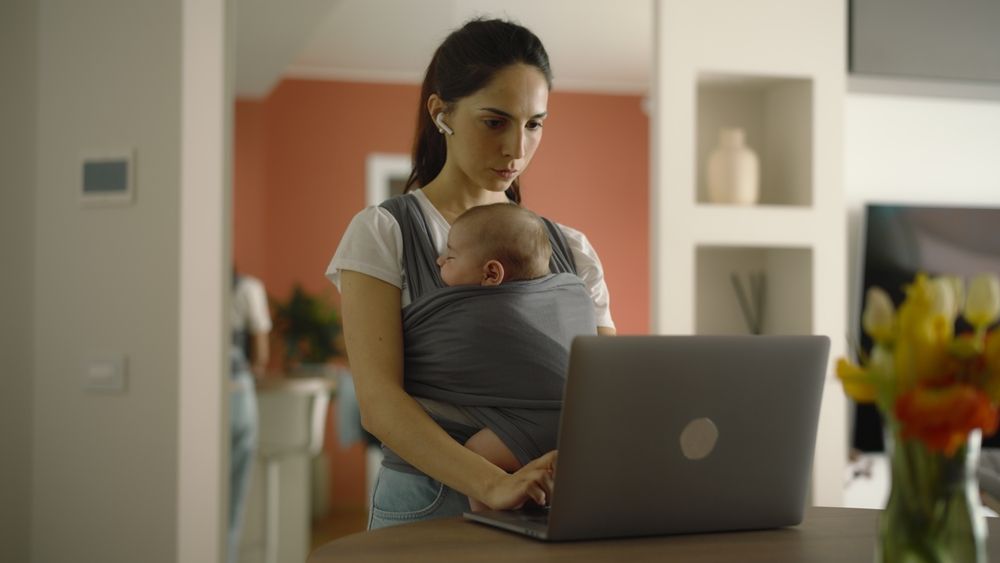 Mutter mit Baby im Tragetuch steht in einer perfekt aufgeräumten Wohnung und arbeitet am Laptop