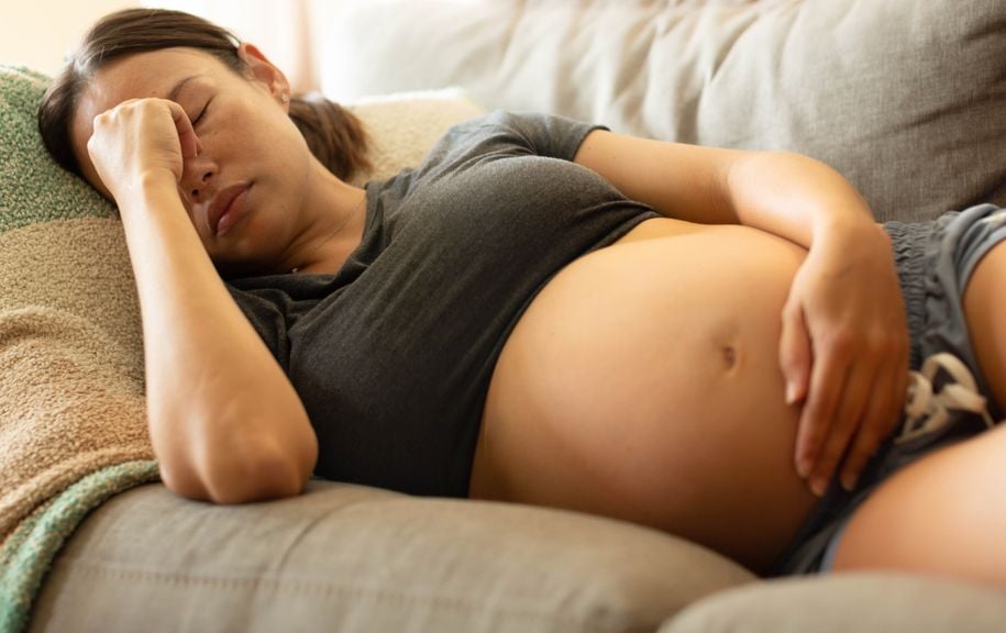 Schwangere liegt auf dem Sofa und hält sich Bauch und Stirne