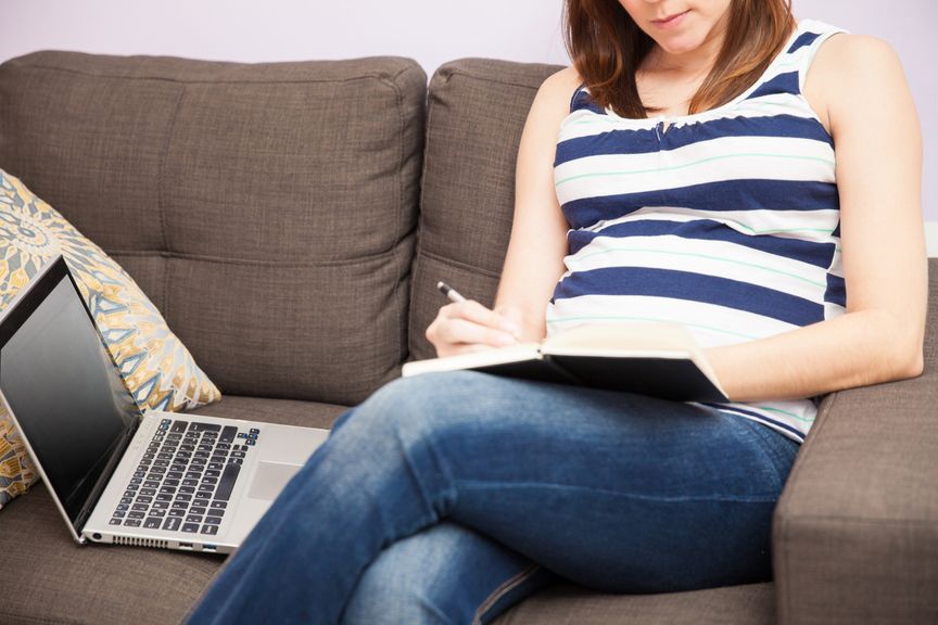 Schwangere sitzt auf dem Sofa mit Laptop und Checkliste oder Tagebuch
