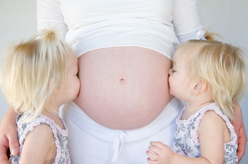Zwillingsmädchen küssen den Babybauch ihrer schwangeren Mutter