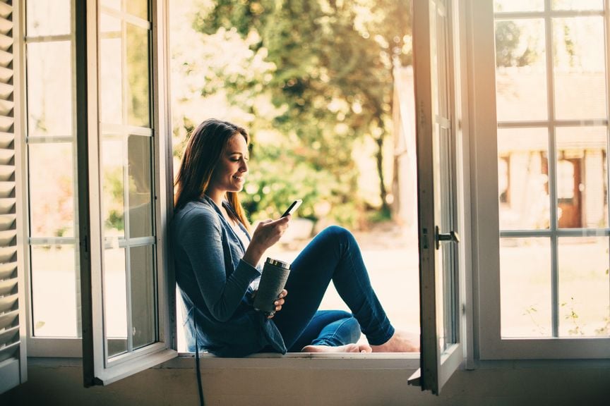 Frau sitzt im Fensterrahmen und schaut in ihr Smartphone