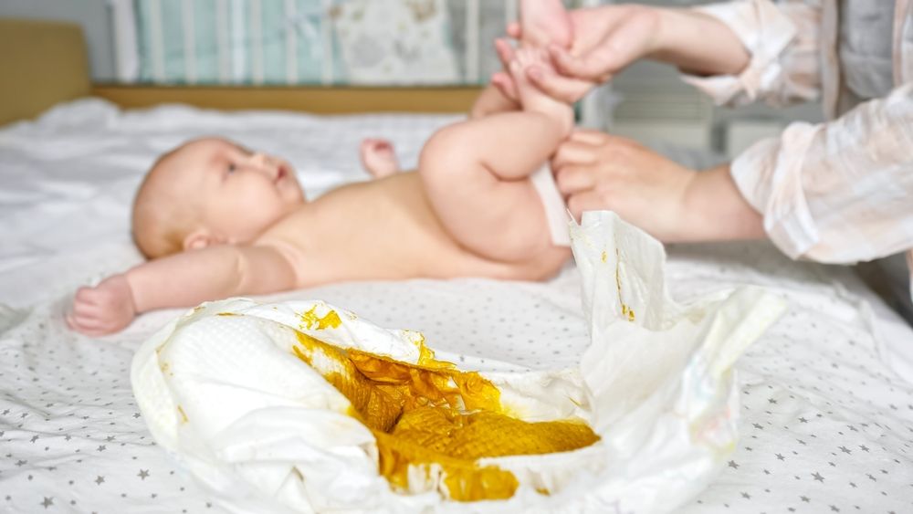 Windel mit Muttermilchstuhl, im Hintergrund eine Mutter, die ihr Baby wickelt.